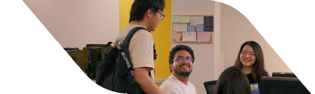 Image 38: A group of students engaged in discussion near computers, with a yellow wall and a bulletin board in the background.
