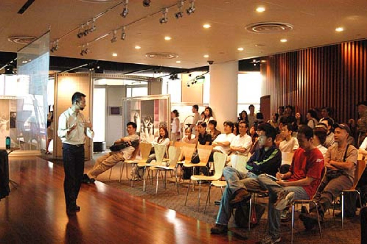 Students and guests seated for a talk at a 3dsense community event, a speaker standing beside display banners of student artwork.