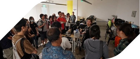 A group of people attentively listens during a classroom presentation with computers and collaborative setups.